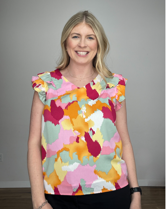 Woman wearing a v-neck sleeveless tank with shoulder ruffles and a watercolor print with dark pink, light pink, seafoam green, white, yellow, and orange colors. 
