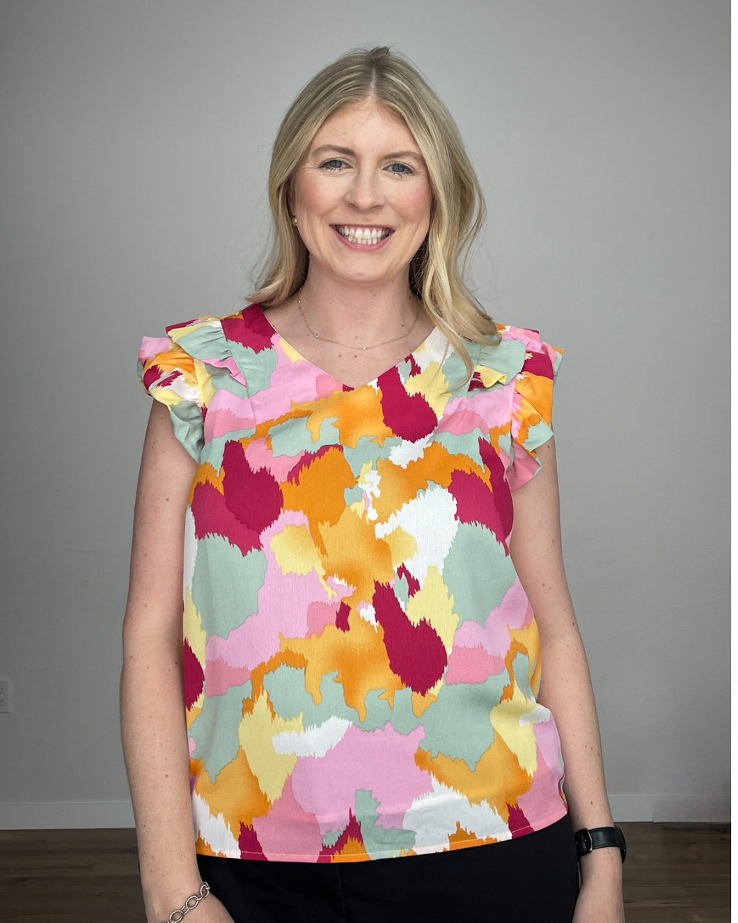 Woman wearing a v-neck sleeveless tank with shoulder ruffles and a watercolor print with dark pink, light pink, seafoam green, white, yellow, and orange colors. 